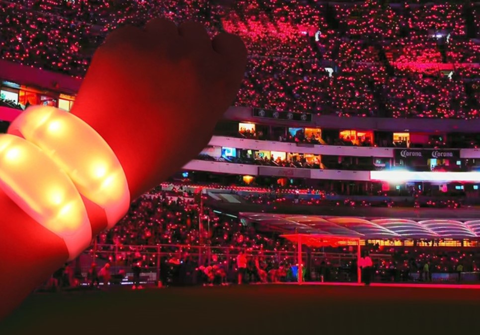 A stadium packed with thousands of fans wearing synchronized glowing concert wristbands creating a coordinated wave of light