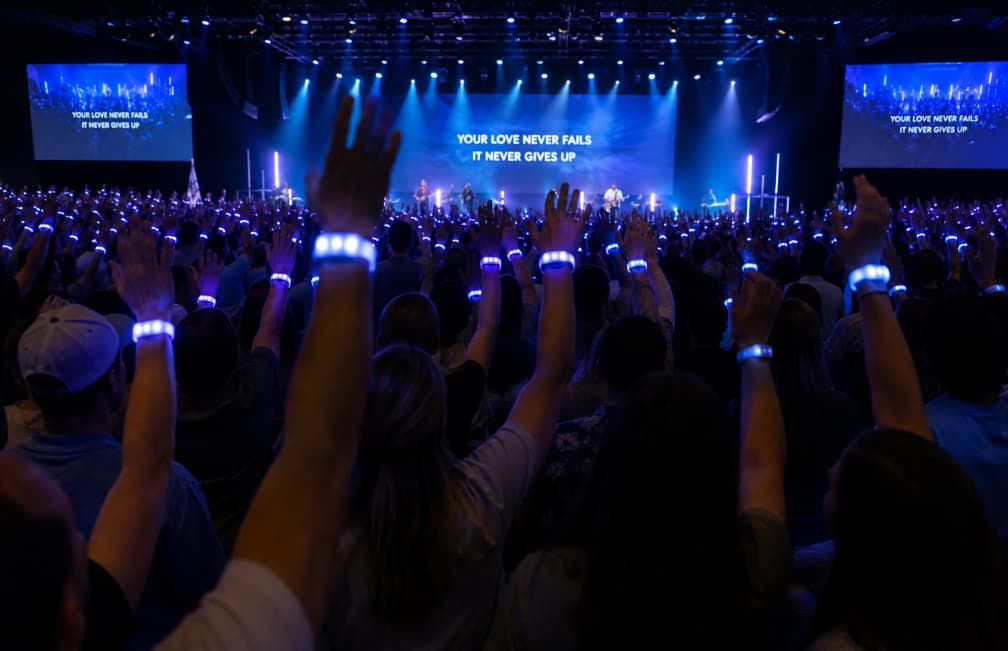 Massive worship arena crowd unified under glowing wireless LED wristbands managed from a central mixing desk