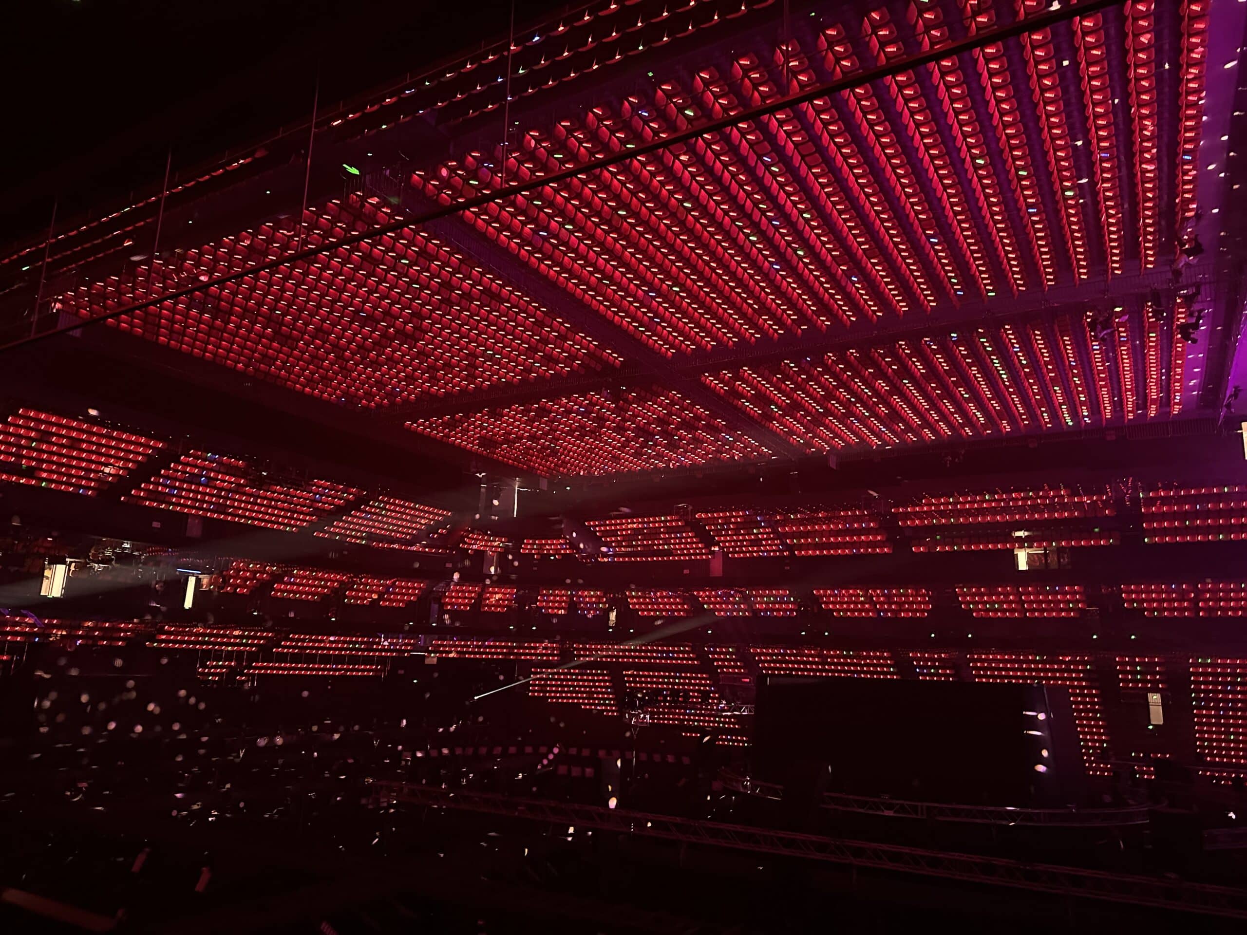 Perspective view from the central lighting control desk looking out toward a massive live concert stage with synchronized color lighting transitions